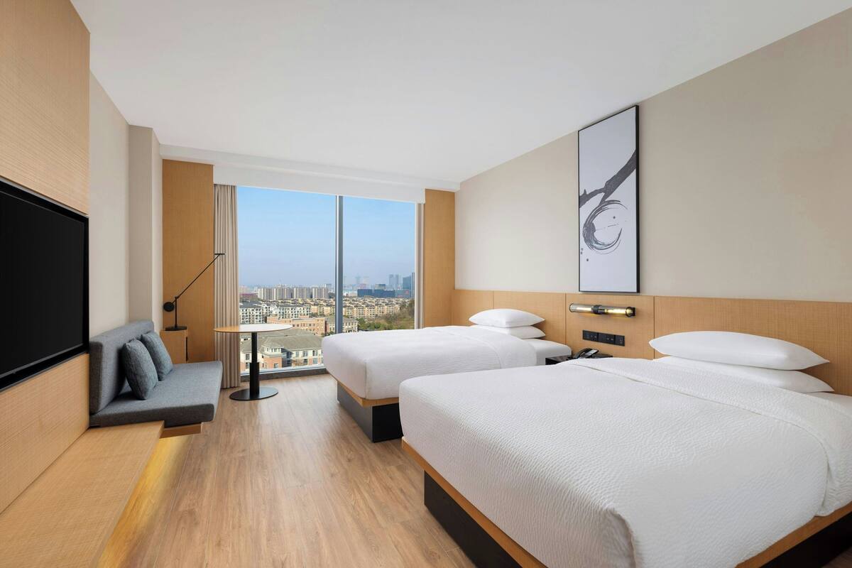 A modern hotel room with two beds, a sofa, a TV, and a large window offering a city view.