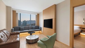 Habitación de hotel moderna con sofá, sillón verde, mesa de centro, televisor de pantalla plana montado en la pared y cama con cabecera.