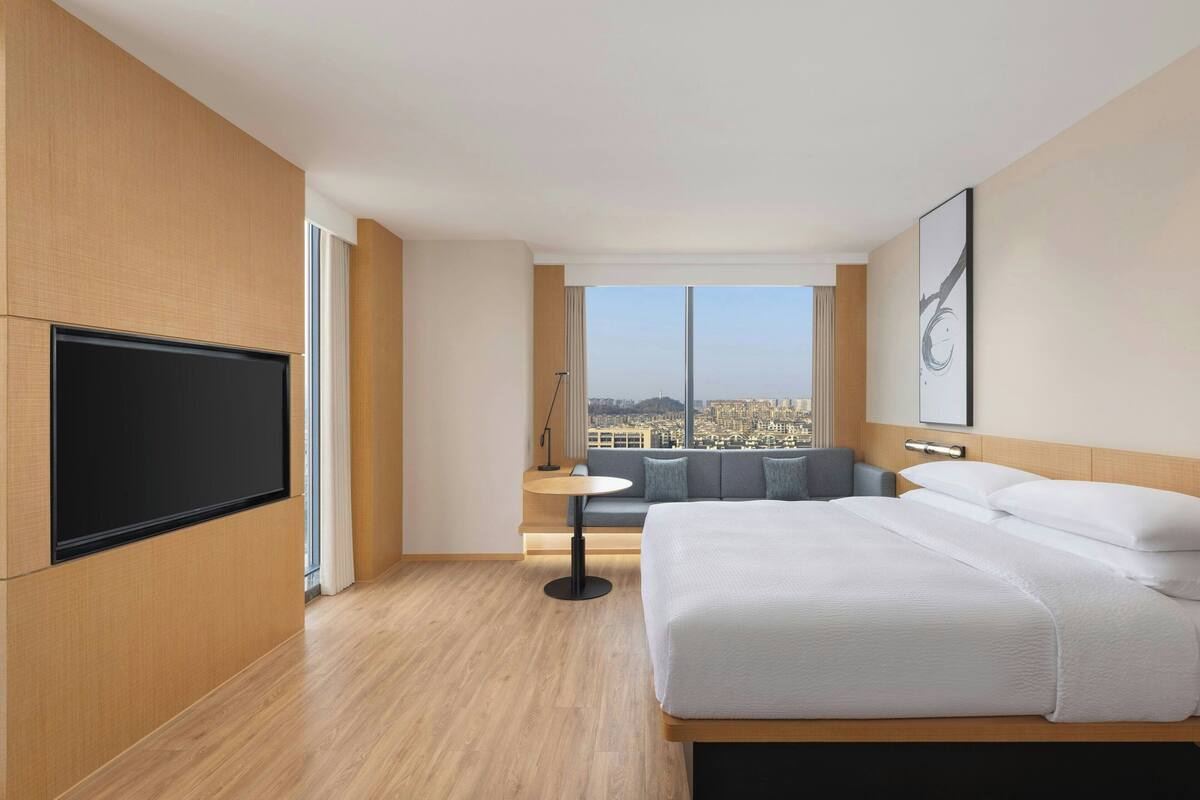 A modern hotel room with a large bed, a flat-screen TV mounted on the wall, a small round table, and a sofa with a city view.