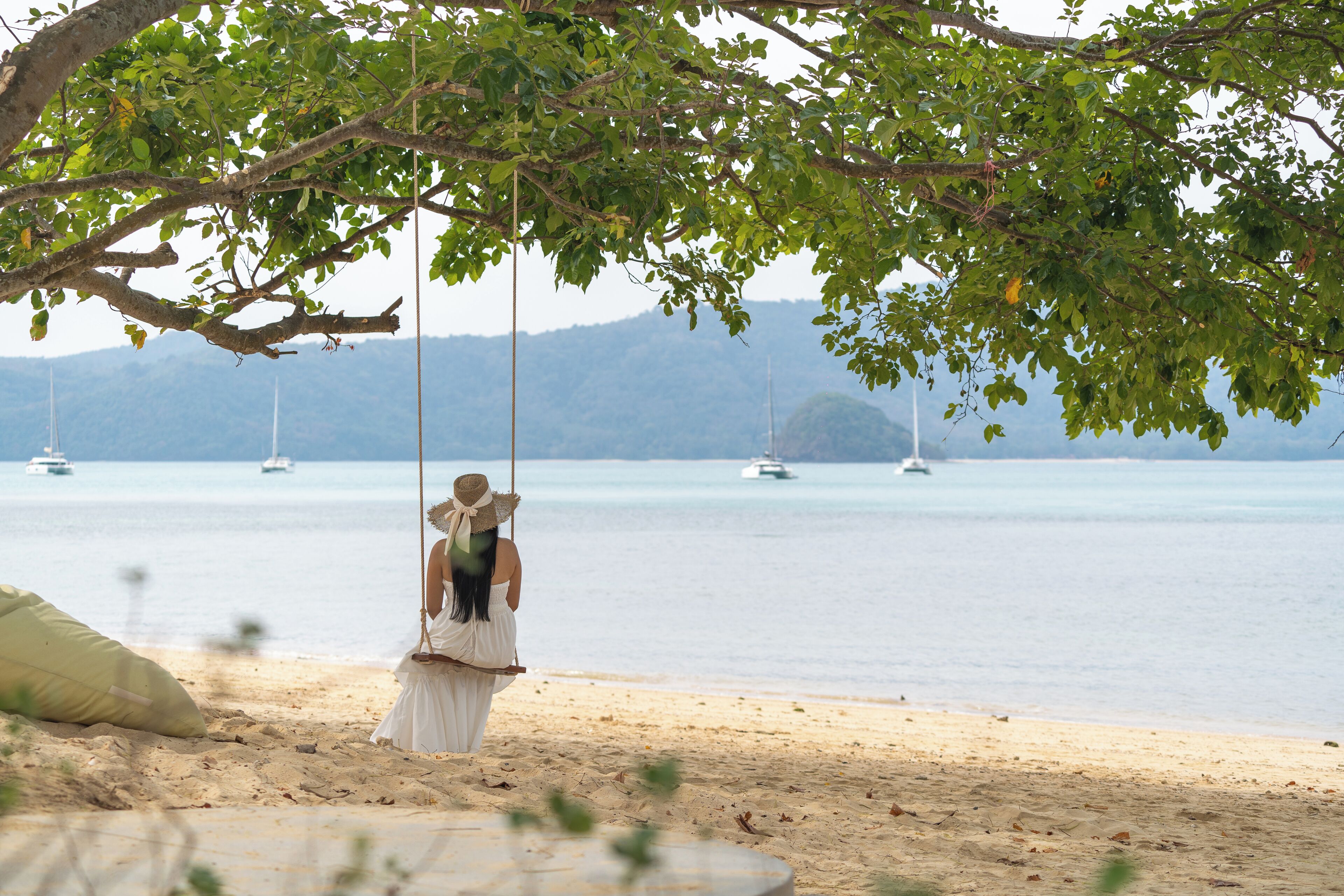 A beach scene with a swing under a tree, a person in a white dress, and boats in the water.