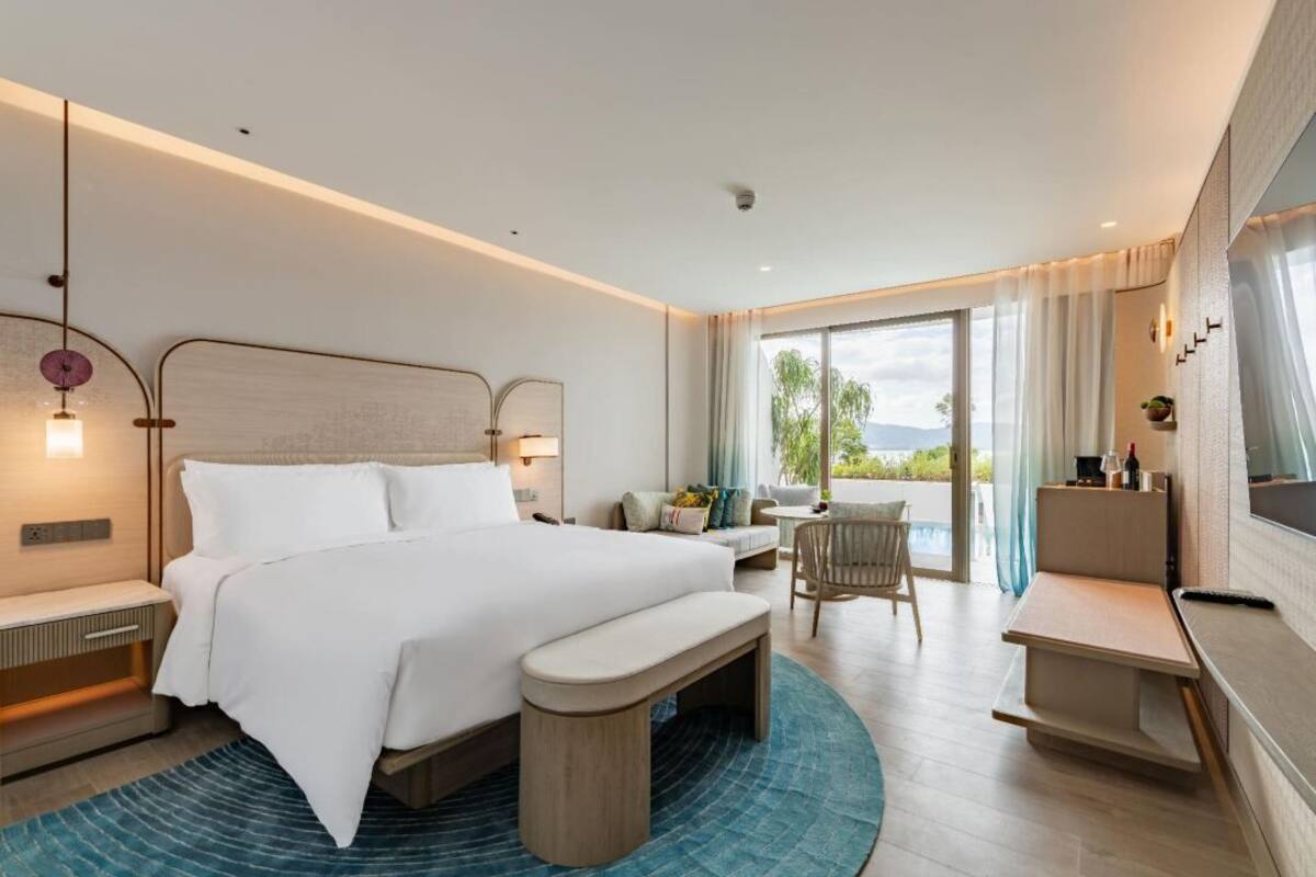 A modern hotel room with a large bed, a desk, a chair, and a view of the outdoors.