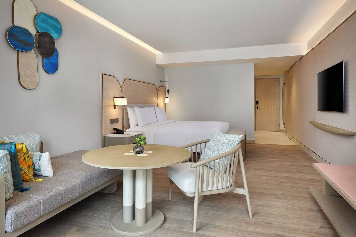 A modern hotel room with a bed, bedside table, lamp, and a round table with a chair.