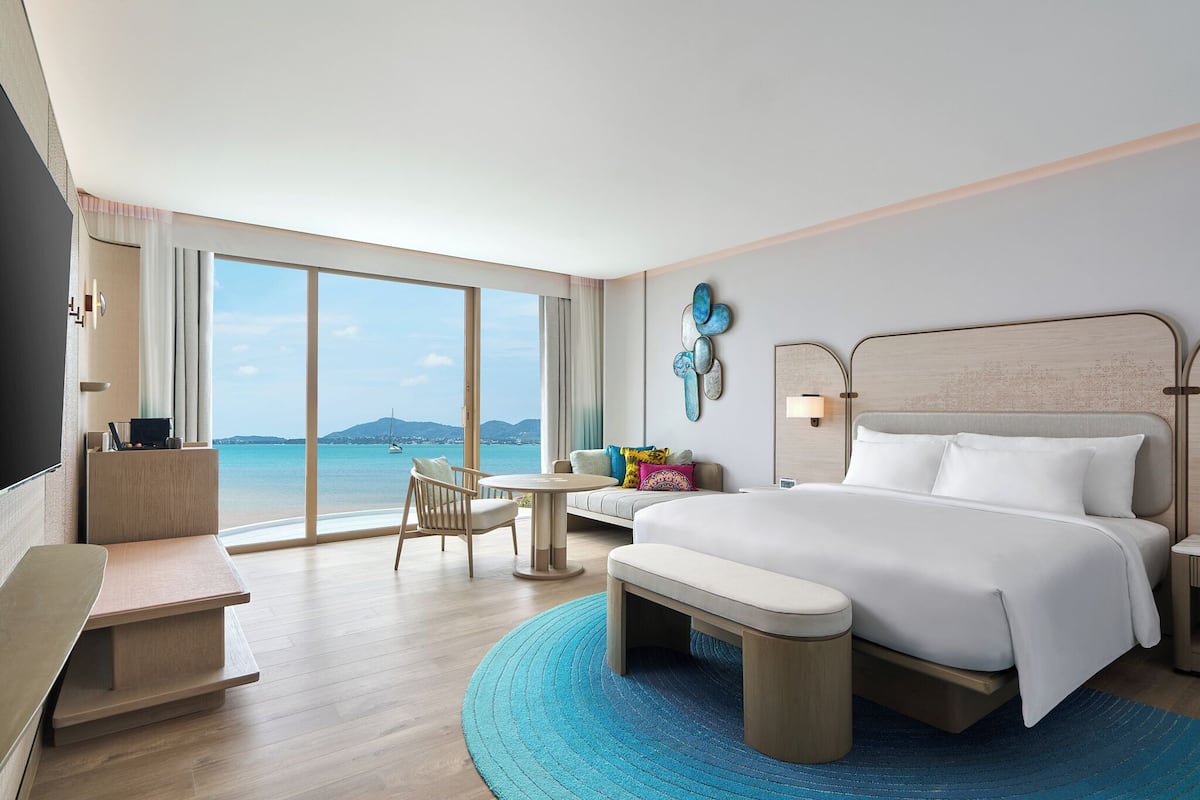 A modern hotel room with a large bed, a desk, a chair, and a view of the sea.