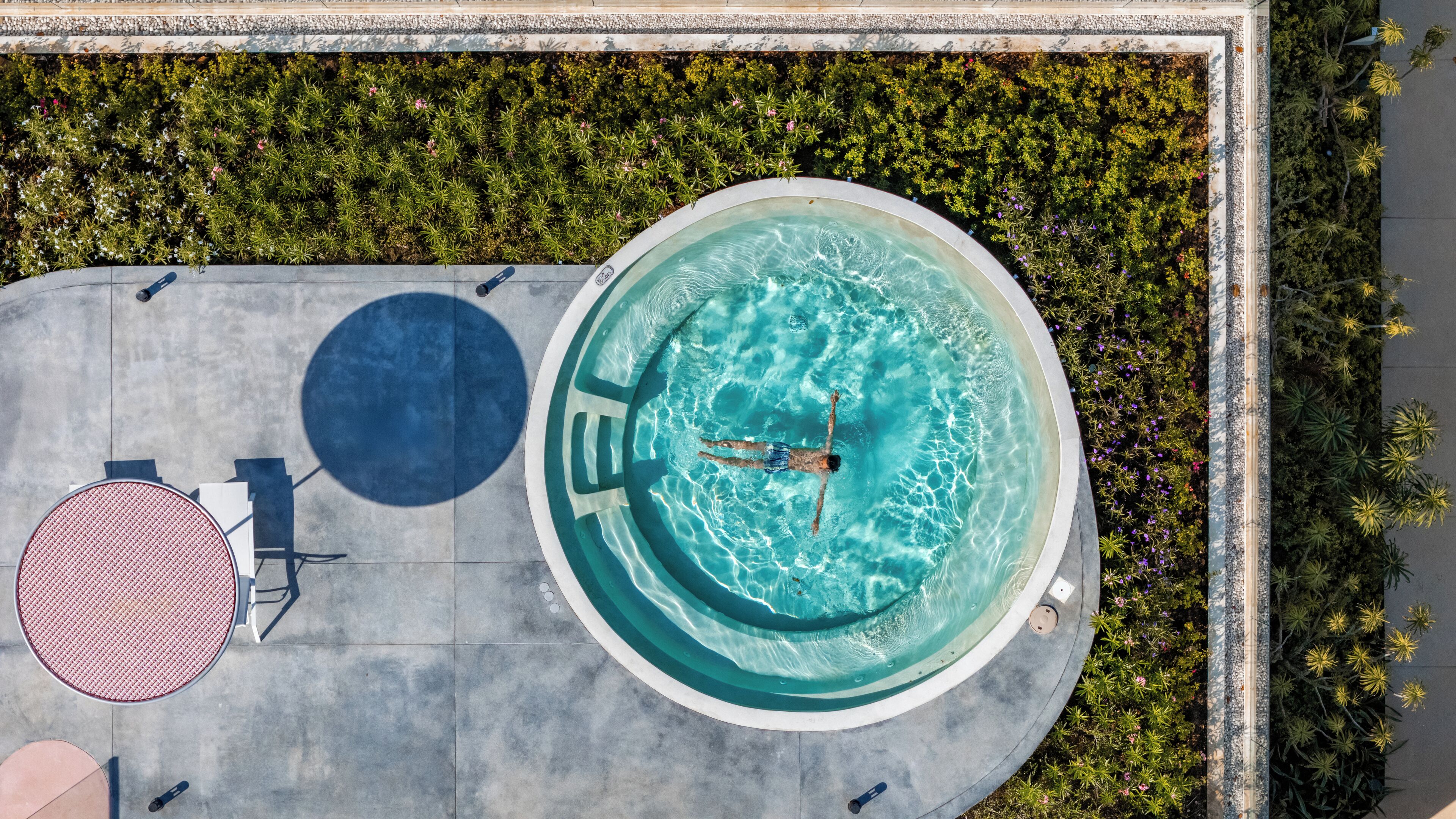 An aerial view of a swimming pool with a person swimming, surrounded by greenery and a tiled deck with seating.