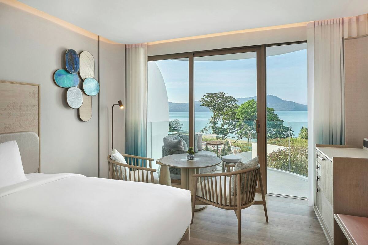 A hotel room with a bed, a round table with chairs, a view of the sea, and a large window.