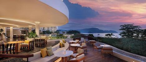 A modern outdoor seating area with round tables and cushioned seating, overlooking a scenic view of the ocean and mountains.