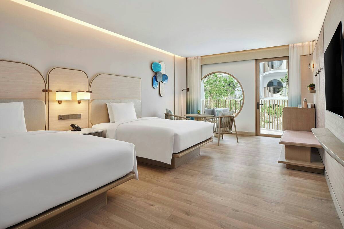 A hotel room with two beds, a TV, a desk, and a balcony with a view of greenery.