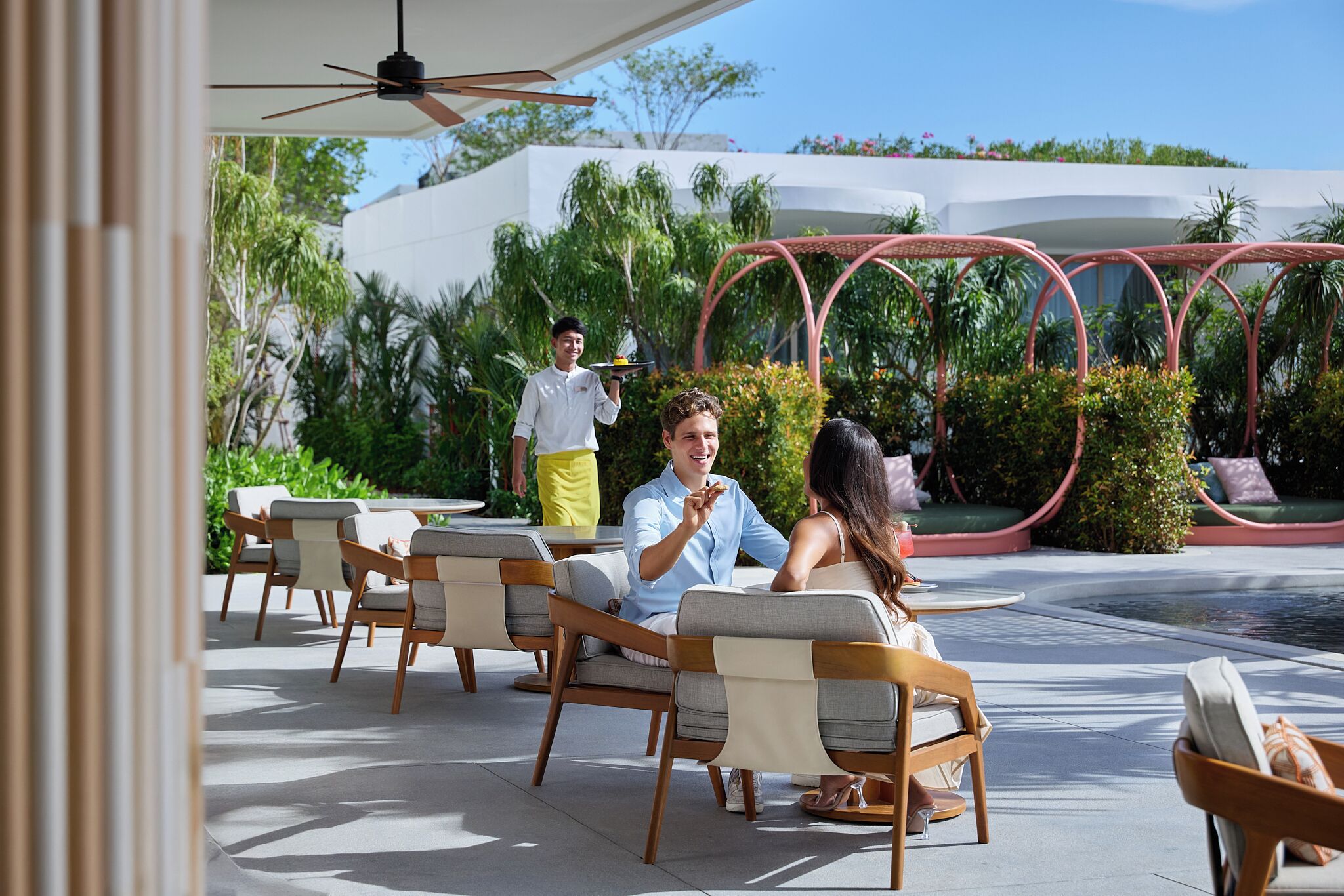 A patio area with outdoor seating, a waiter serving, and a pool in the background.