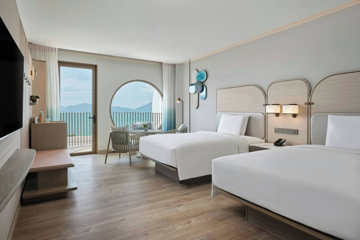 A hotel room with two beds, a desk, a chair, and a balcony with a view of the mountains.
