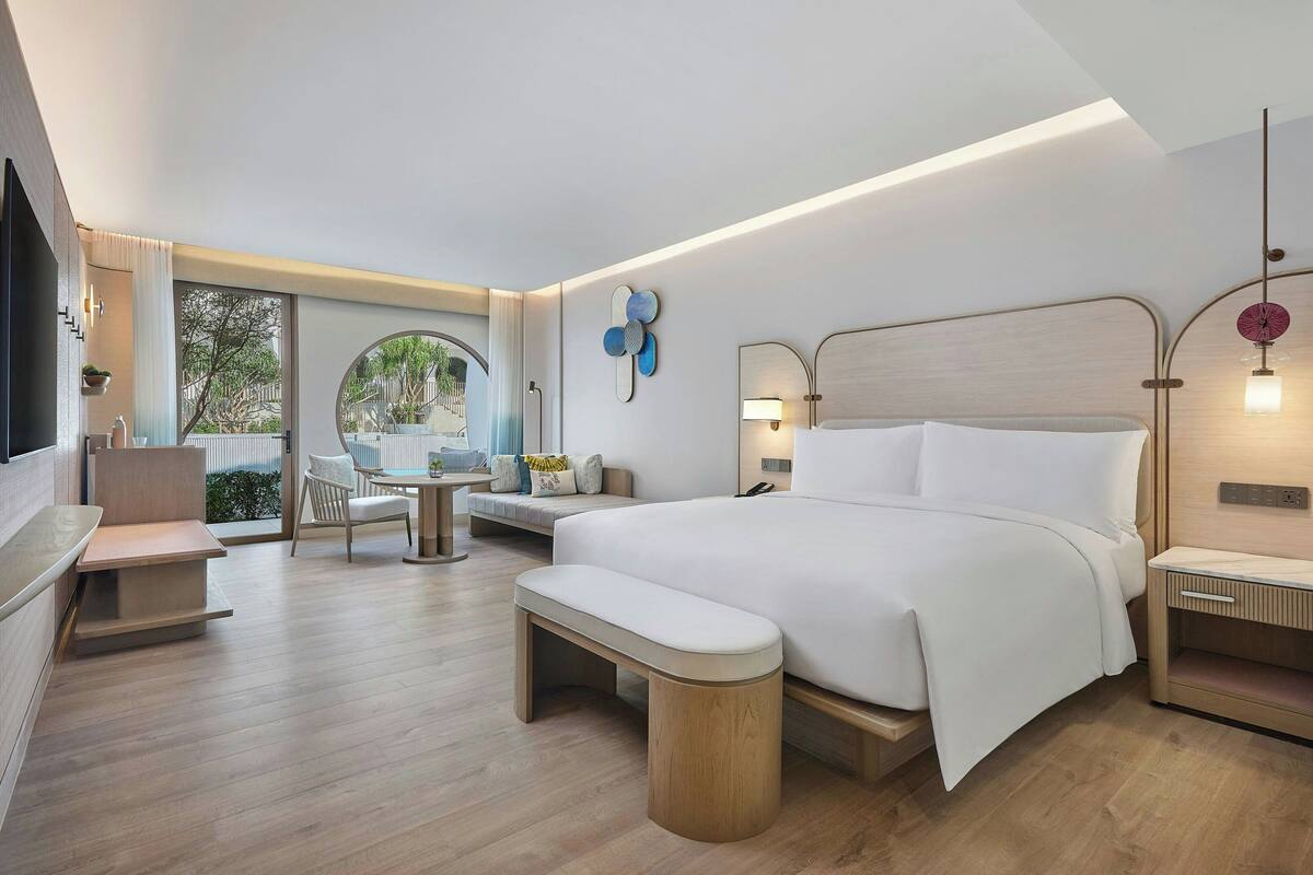 A modern hotel room with a large bed, a bench, a desk, and a view of greenery outside.