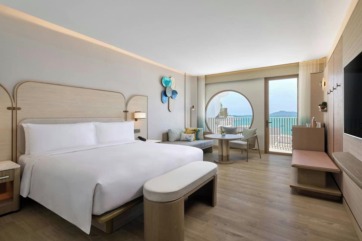 A modern hotel room with a large bed, a small seating area, and a balcony with a view.