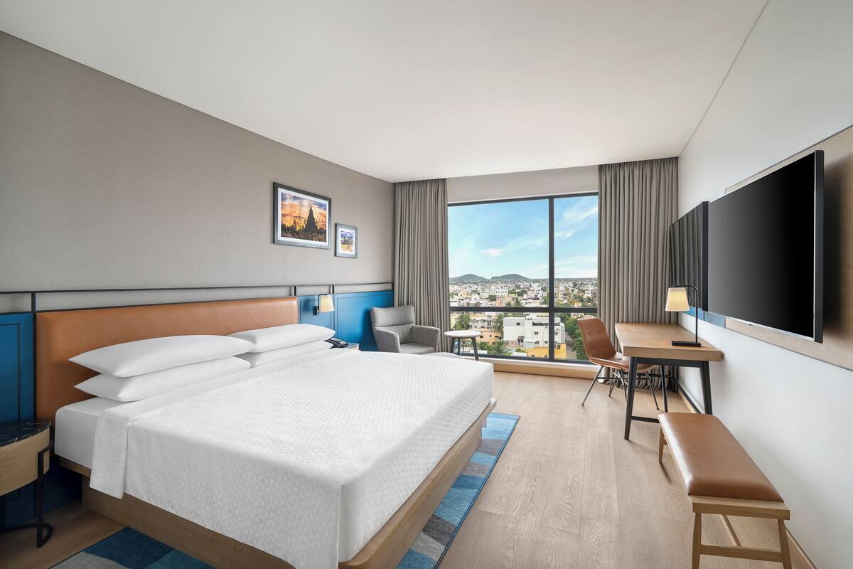 Habitación de hotel con una cama grande, un escritorio, una silla, un televisor y vistas a la ciudad.