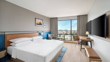 A hotel room with a large bed, a desk, a chair, a TV, and a view of the city.