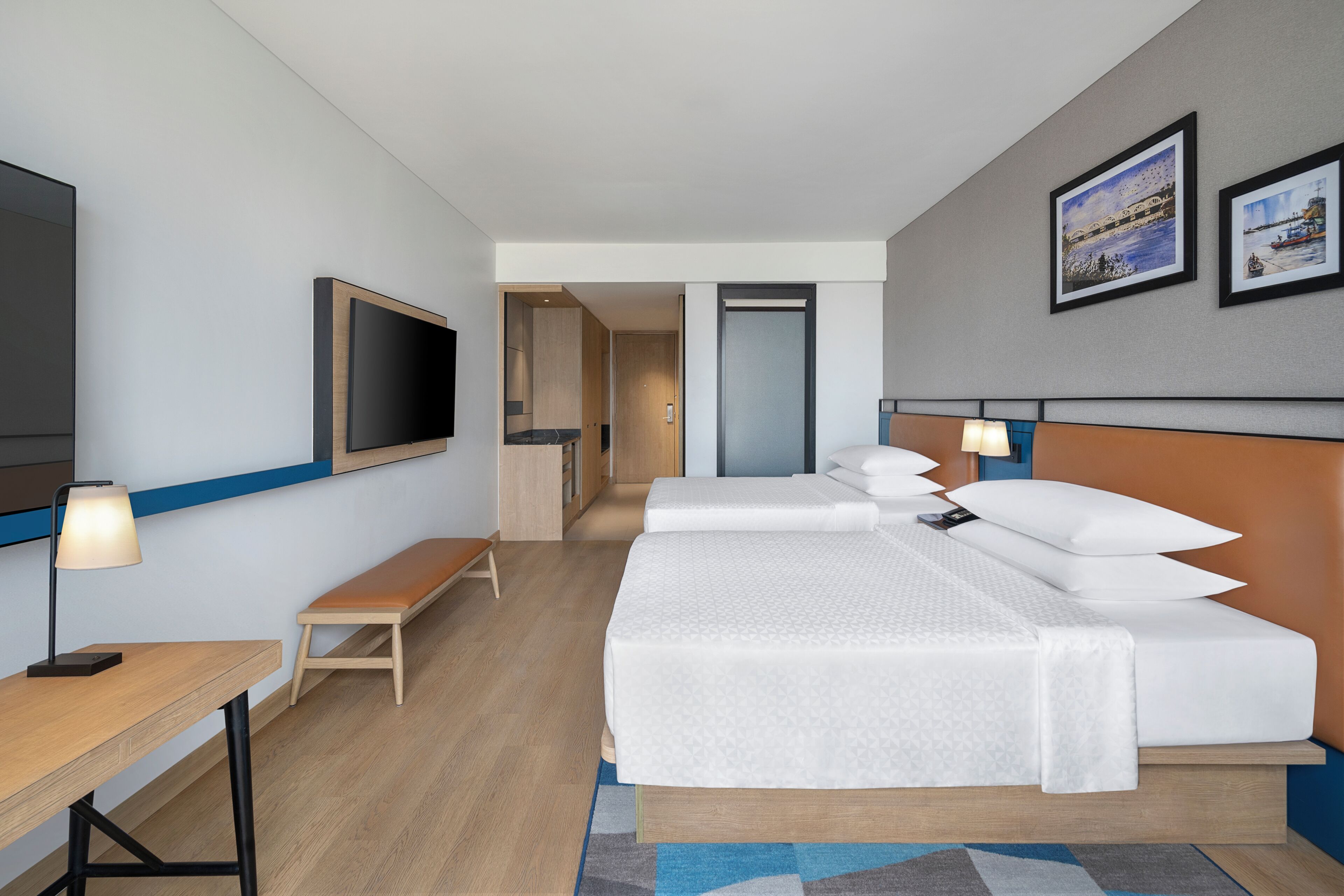 Habitación de hotel con cama, mesitas de noche, televisor, banco y puerta que da al baño.