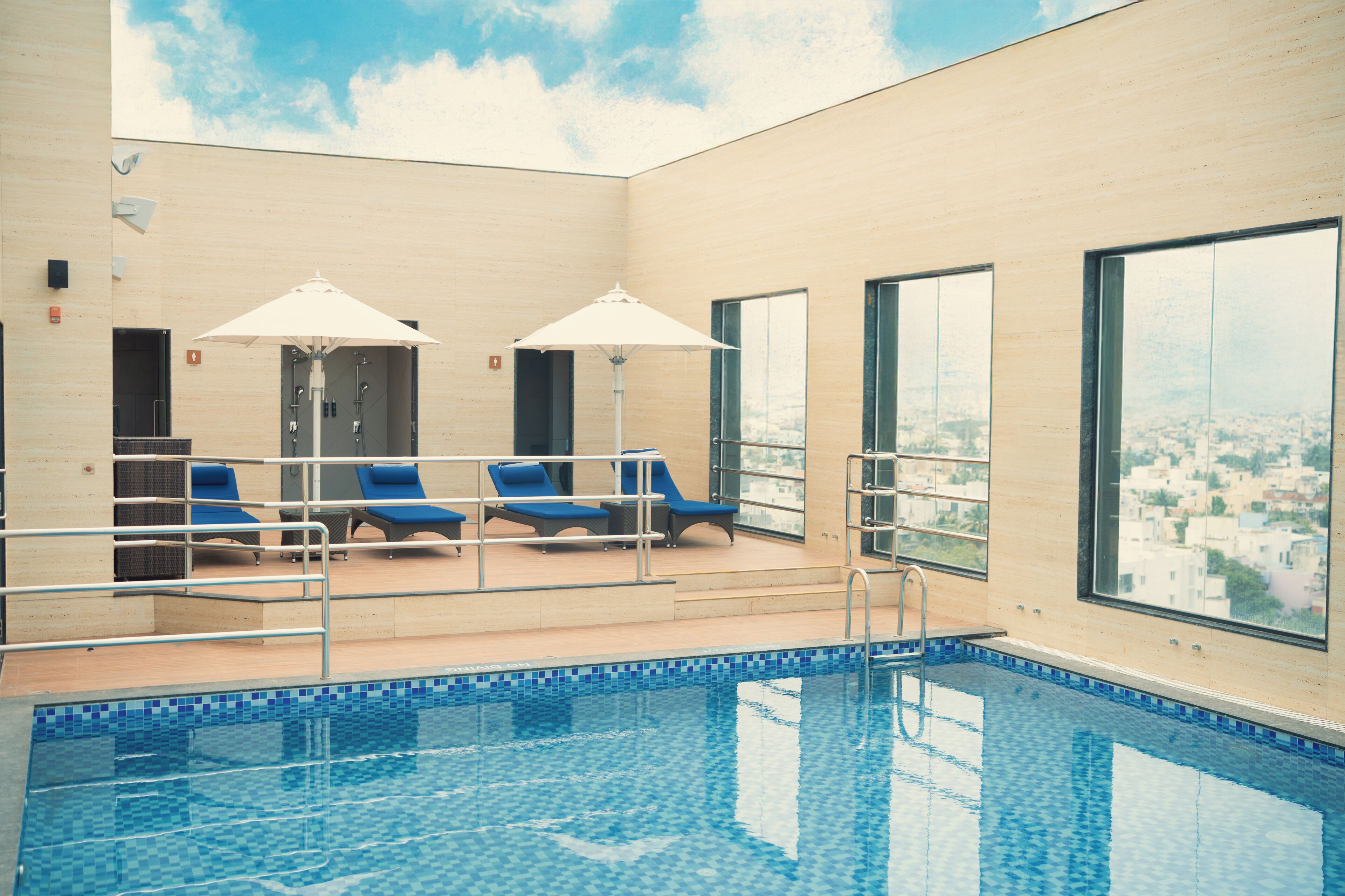 An indoor swimming pool with blue tiled flooring, a pool ladder, and blue lounge chairs under white umbrellas.
