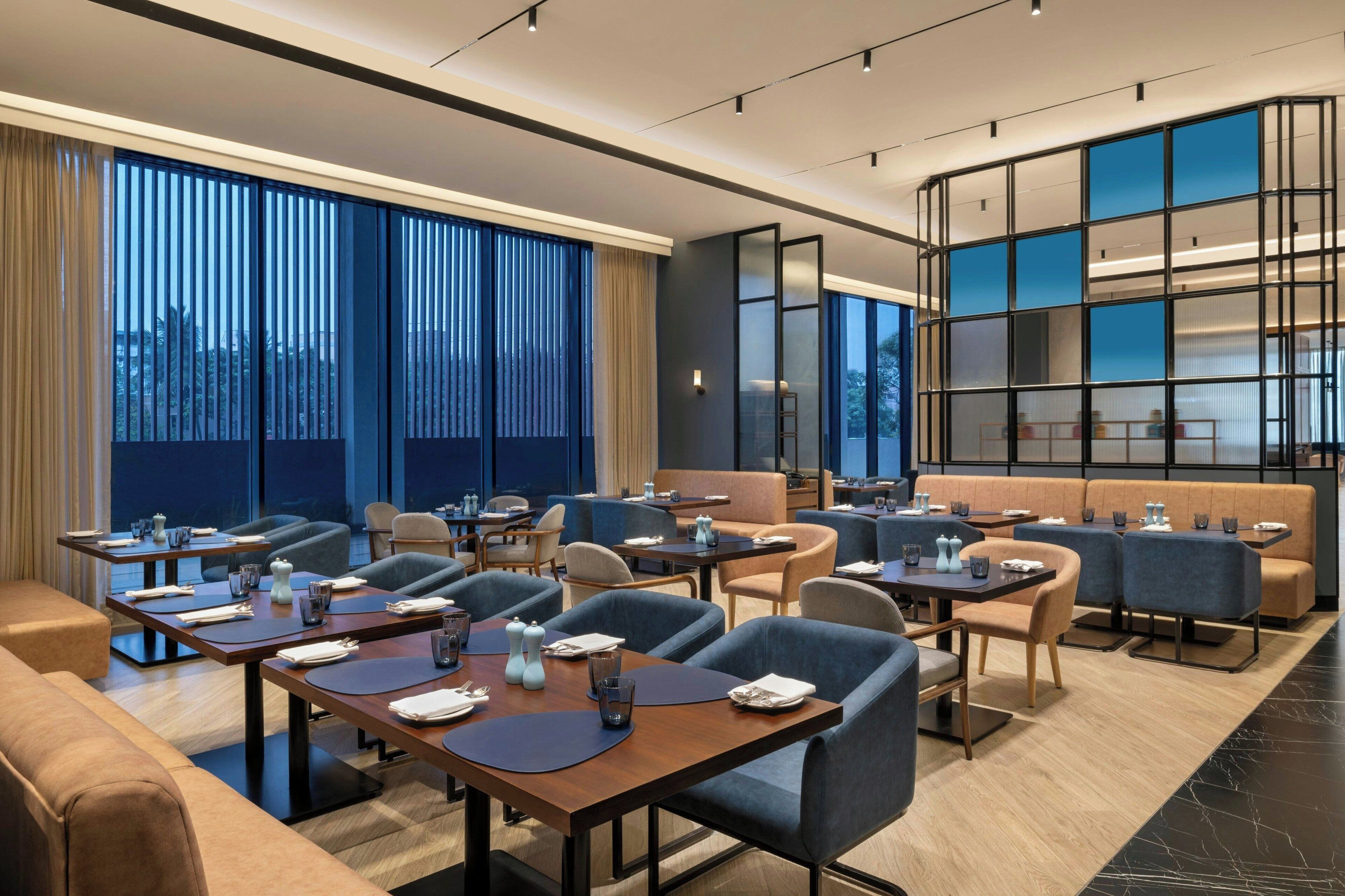 A modern hotel dining area with multiple tables and chairs, large windows with curtains, and a seating area with a sofa and a bar.