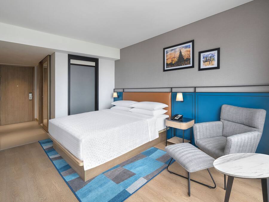 A hotel room with a bed, a chair, a small table, a desk, and a door.