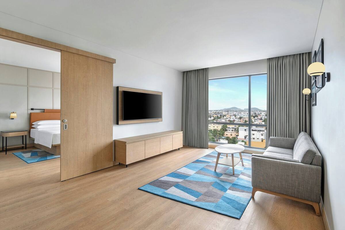 Una habitación de hotel moderna con un ventanal grande, un televisor de pantalla plana en la pared, un sofá, una mesita redonda y un mueble de madera.