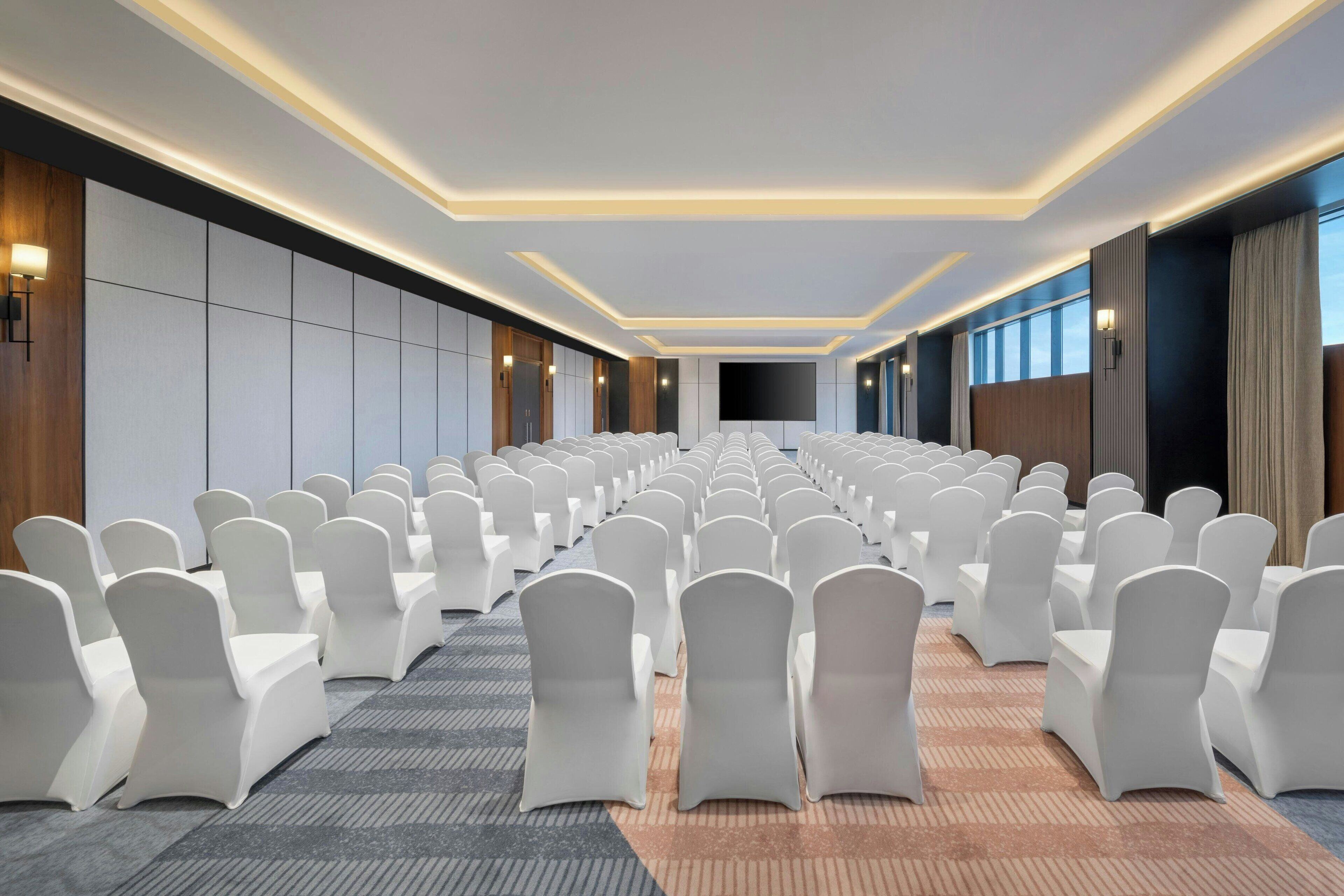 A conference room with rows of white chairs and tables, a carpeted floor, and a modern ceiling design.