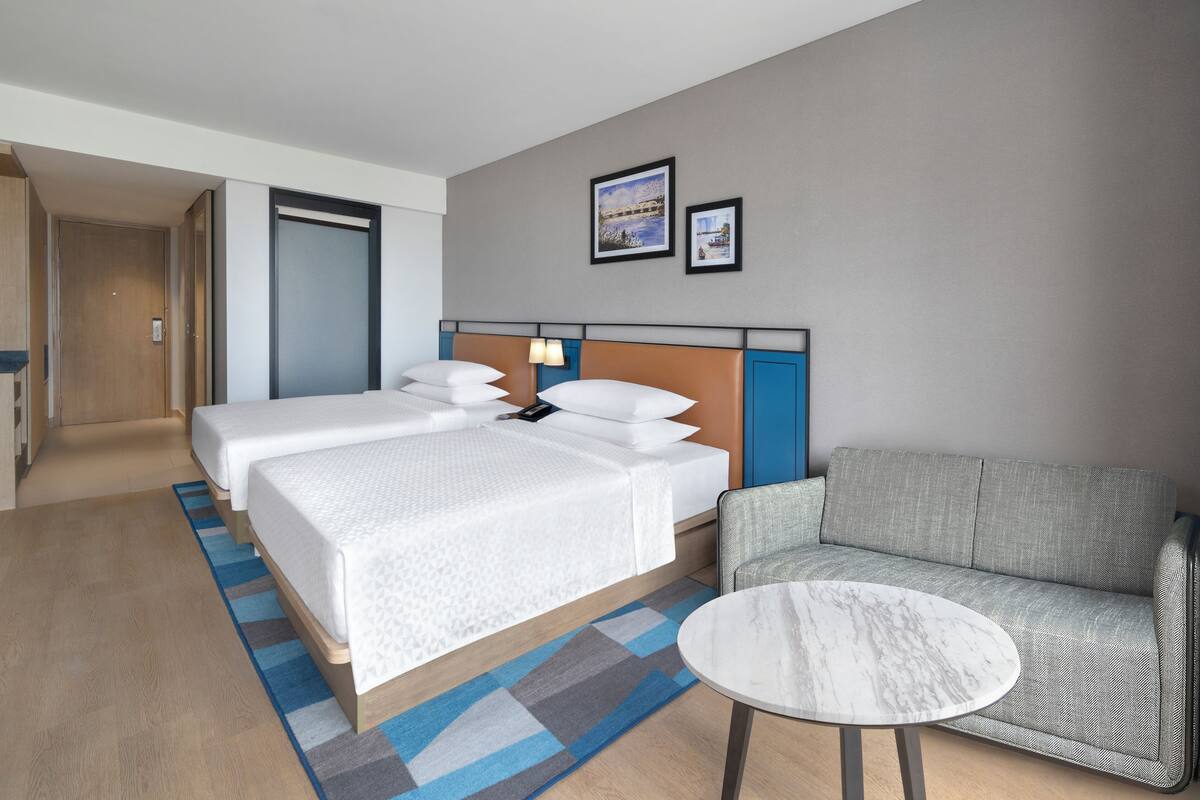 Habitación de hotel con dos camas, un sofá, una mesa redonda y una puerta que da a otra habitación.