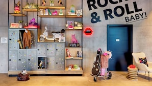 Une pièce avec une grande enseigne murale sur laquelle on peut lire : « CE N’EST QUE DU ROCK AND ROLL, MON CHÉRI ! », des étagères remplies de divers objets, une guitare et une chaise blanche.