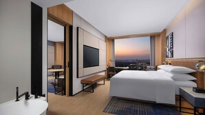 Una habitación de hotel moderna con una cama grande, un televisor de pantalla plana, un escritorio con una silla y vistas a la ciudad al atardecer.