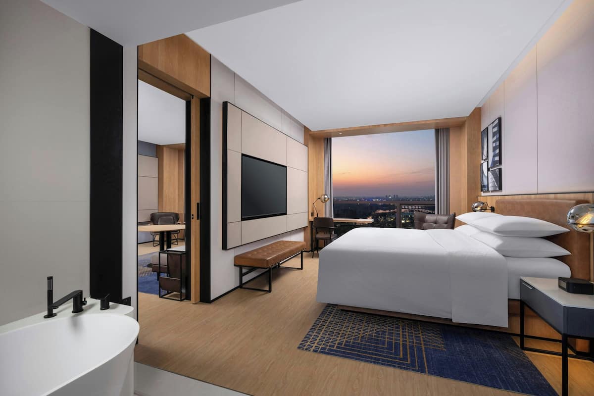 Une chambre d’hôtel moderne dotée d’un grand lit, d’un téléviseur à écran plat, d’un bureau avec une chaise et offrant une vue sur la ville au coucher du soleil.