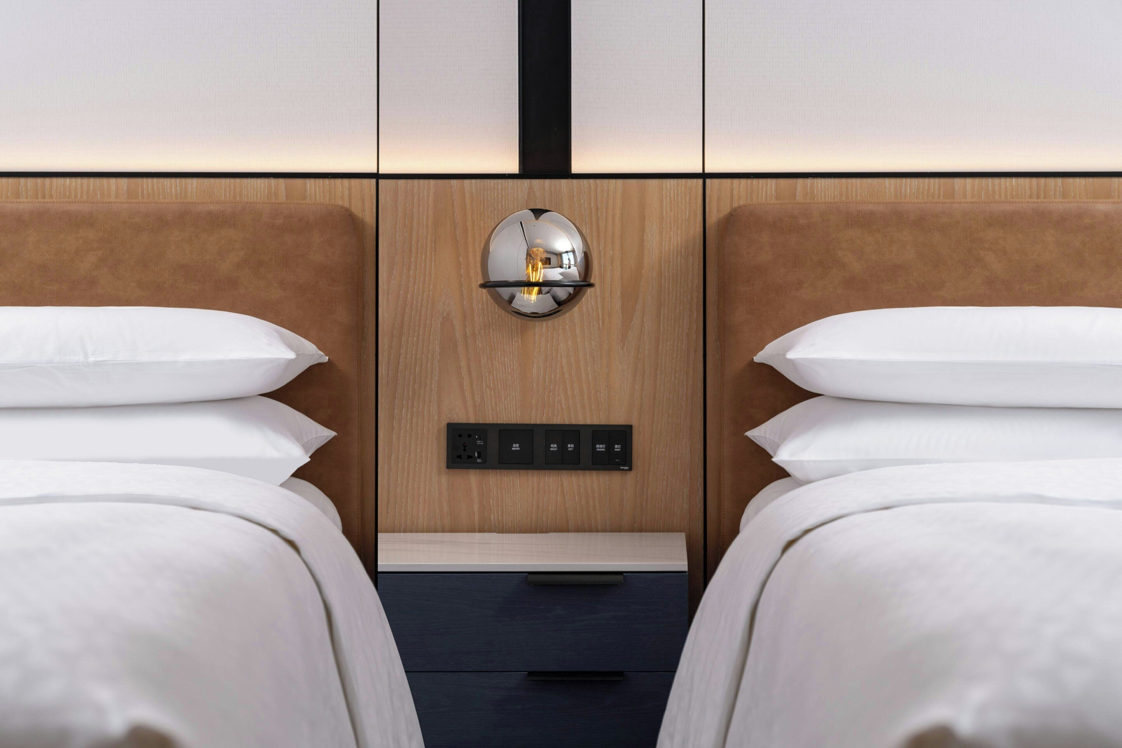 A hotel room with two beds, a wooden headboard, a wall-mounted light fixture, and a control panel.