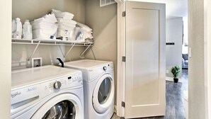 Pesuhuoneessa on kaksi valkoista Whirlpool-merkkiä olevaa pesukonetta, hyllyllä on siististi taiteltuja pyyhkeitä ja ovi johtaa toiseen huoneeseen.