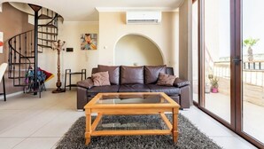 Un salon moderne avec un canapé en cuir marron, une table basse en verre, un escalier en colimaçon et une grande fenêtre donnant sur l’extérieur.