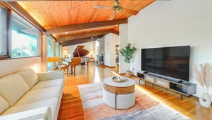 Ein geräumiges Wohnzimmer mit großem Fernseher, Holzbalkendecke, einem Klavier und einer Couch.