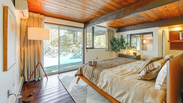 Ein Schlafzimmer mit Holzbalkendecke, einem großen Bett mit beigefarbener Bettdecke, einem Nachttisch mit Lampe und einer Glastür zum Balkon.