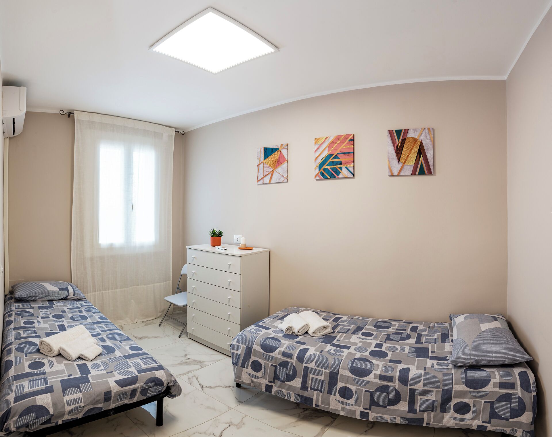 A bedroom with two beds, a desk, and a chair. There are three wall-mounted artworks and a ceiling light.