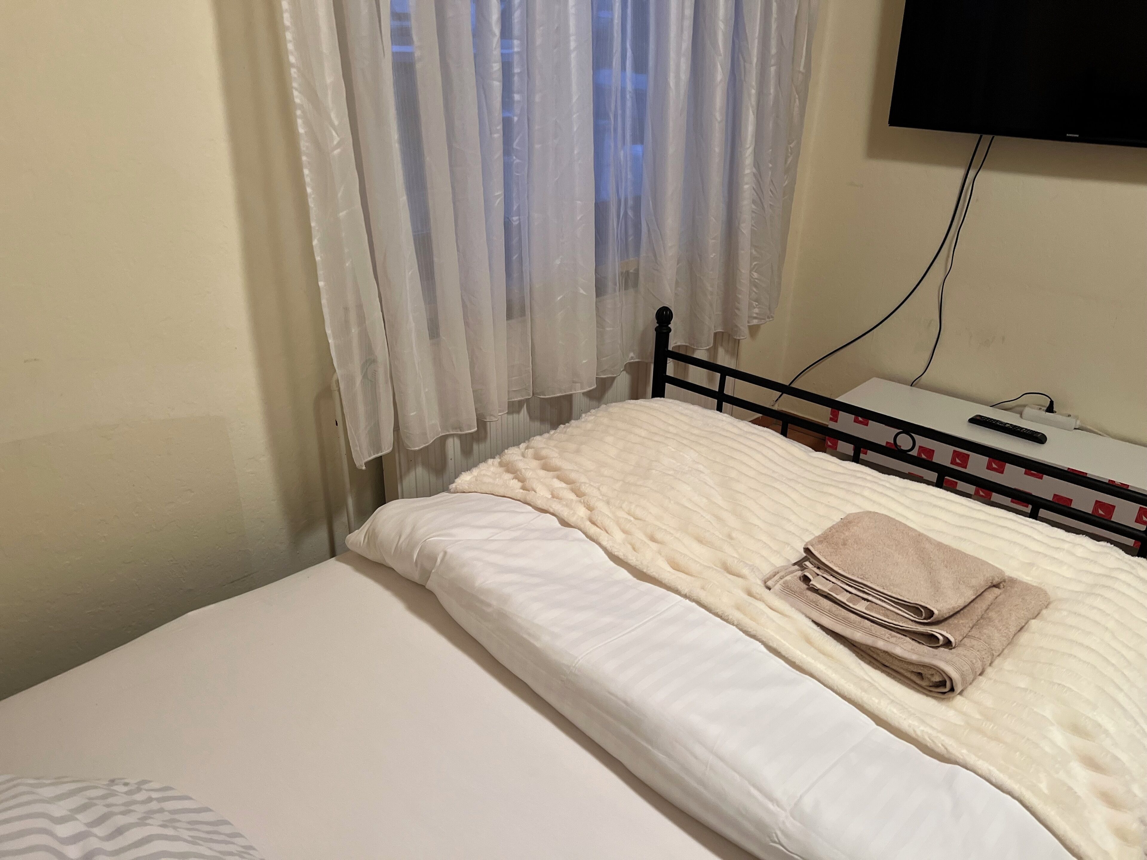 Een enkel bed met een witte dekbedovertrek en een beige handdoek, een aan de muur bevestigde televisie en een raam met transparante gordijnen.