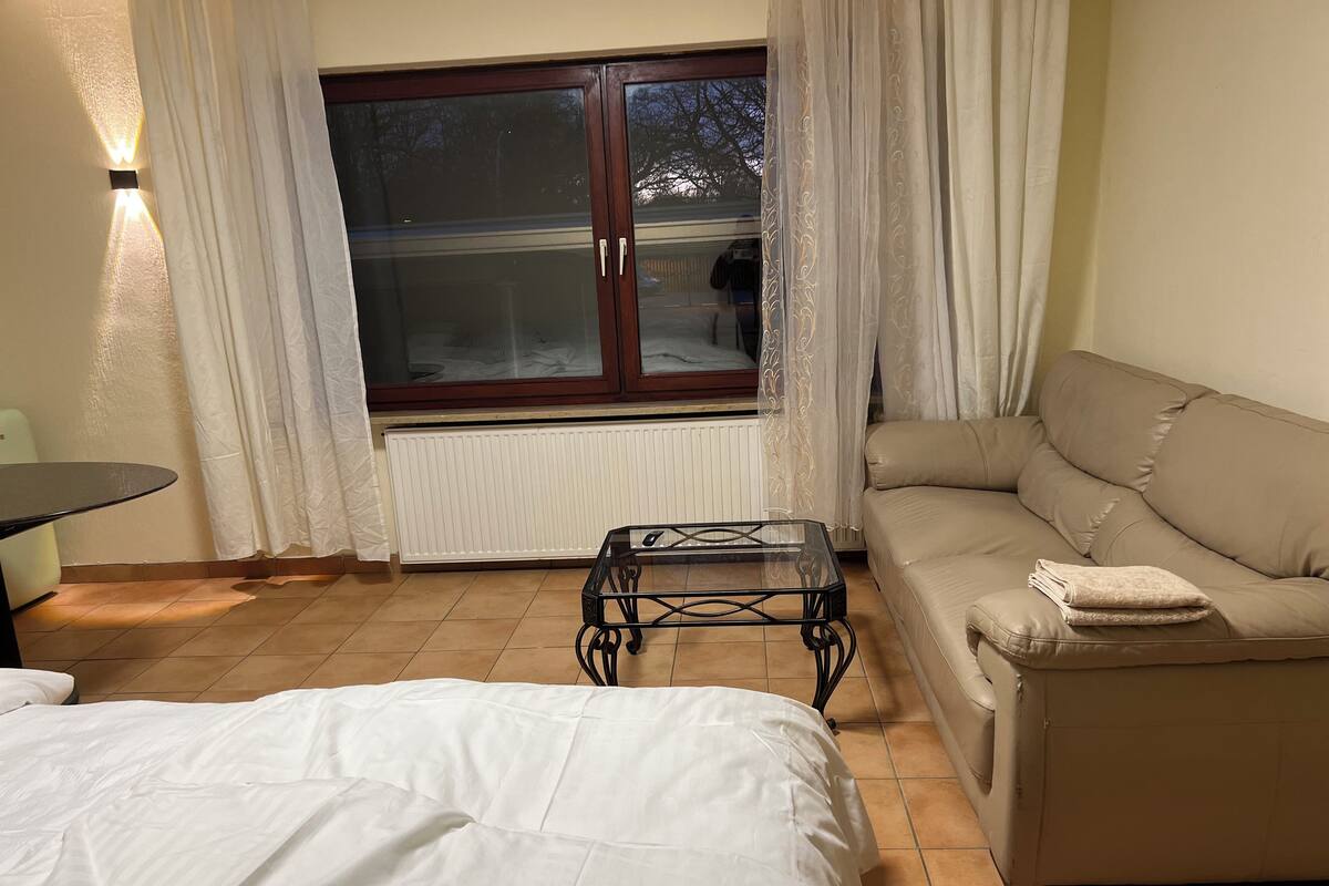 A bedroom with a bed, a small table, a sofa, and a window with curtains.