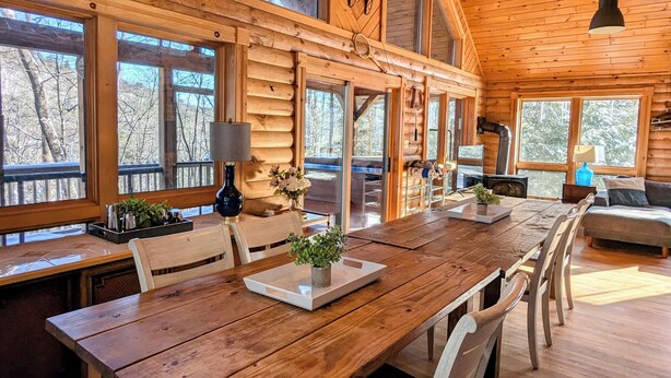 A spacious wooden cabin with a large dining table, multiple windows, and a unique antler chandelier.