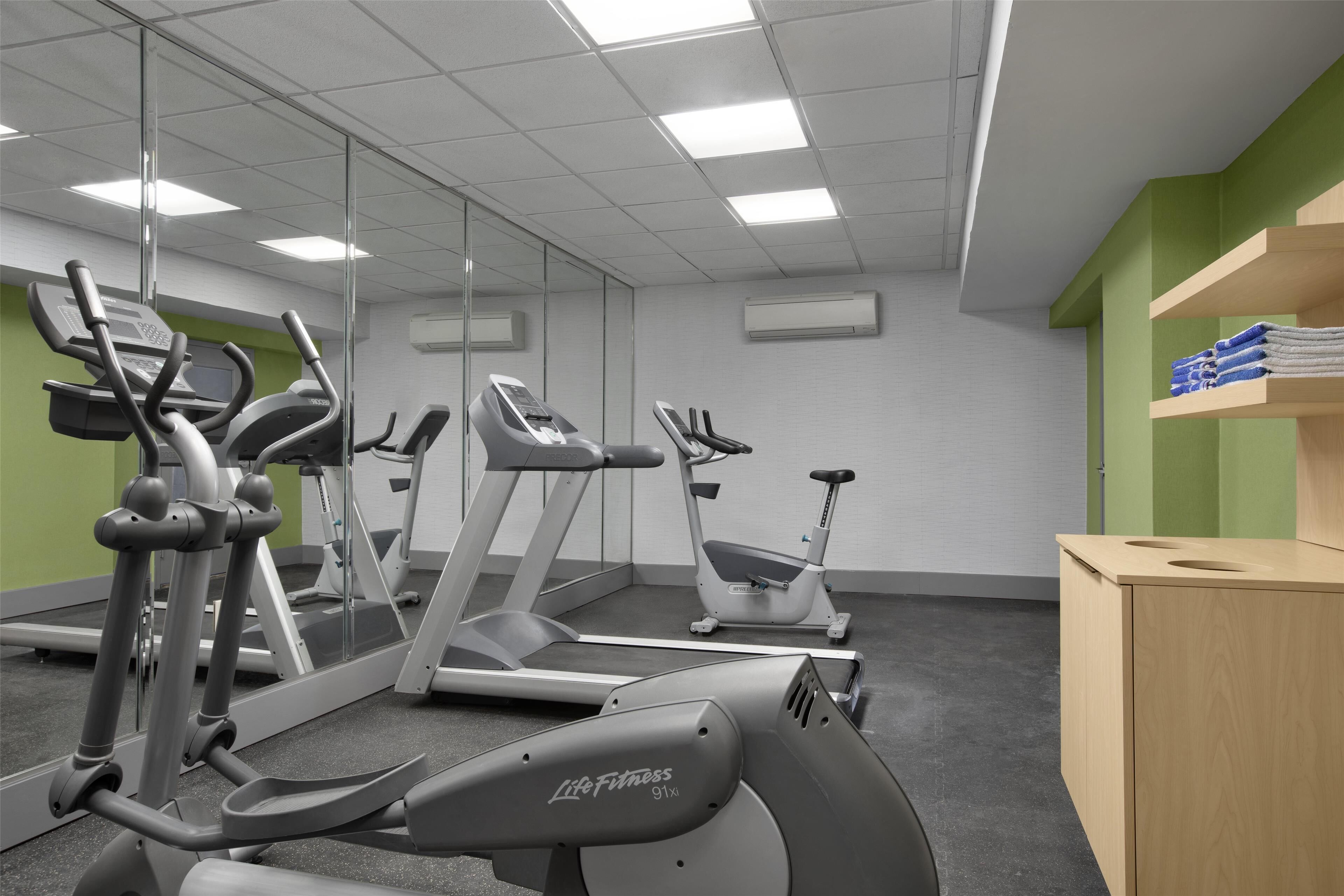 Une salle de sport équipée de machines de cardio, de miroirs et d’un meuble en bois contenant des serviettes.