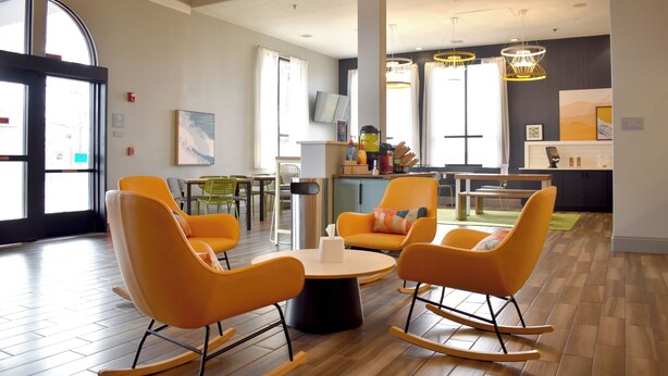 A modern living room with orange chairs, a round table, and a kitchen area in the background. - Spark By Hilton Albuquerque North I-25 (Albuquerque)