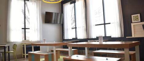 Uma sala de reuniões moderna com várias mesas e cadeiras, amplas janelas com cortinas e uma TV de tela plana na parede.