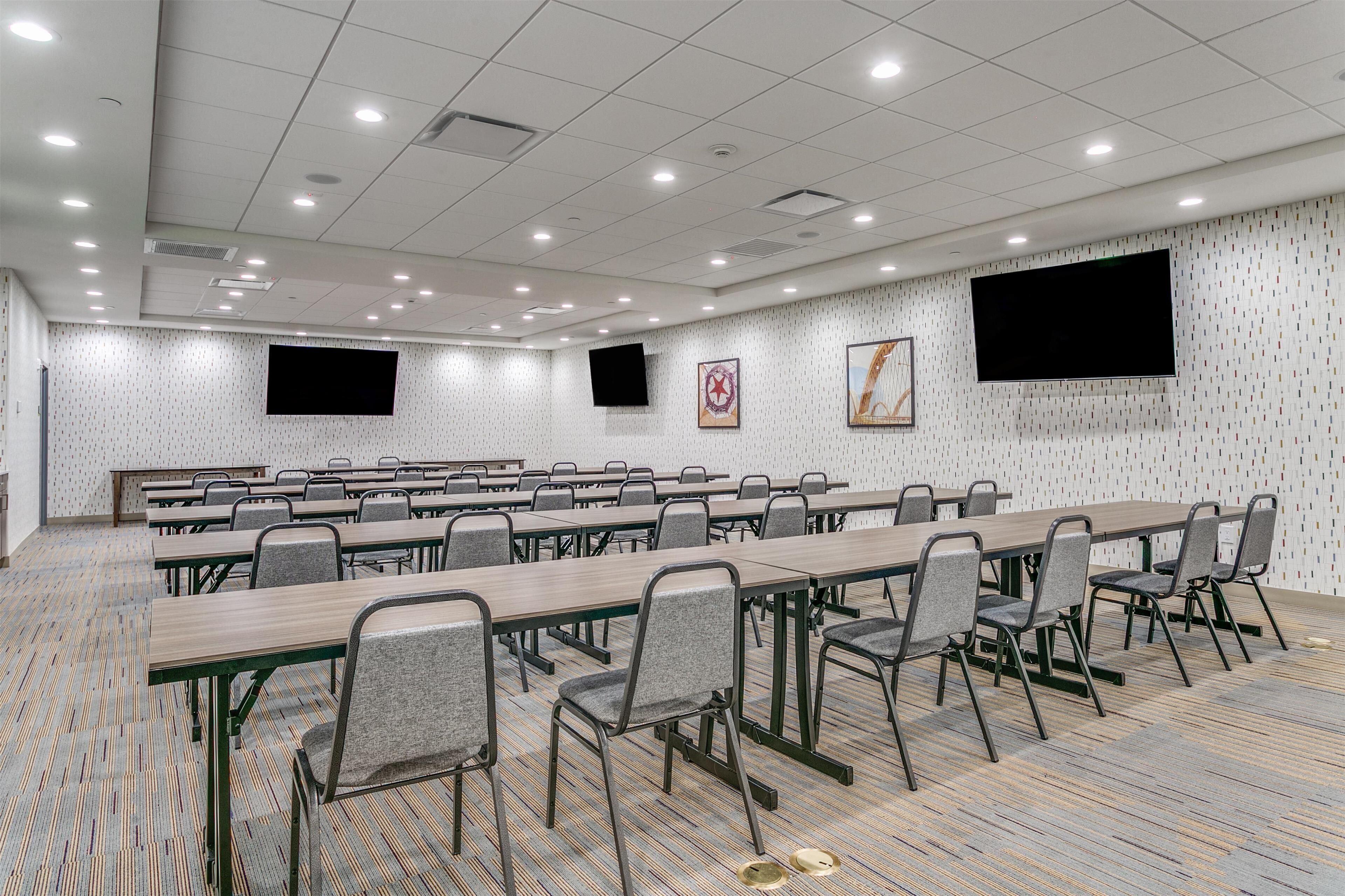 A conference room with multiple tables and chairs, a flat-screen TV mounted on the wall, and patterned wallpaper.