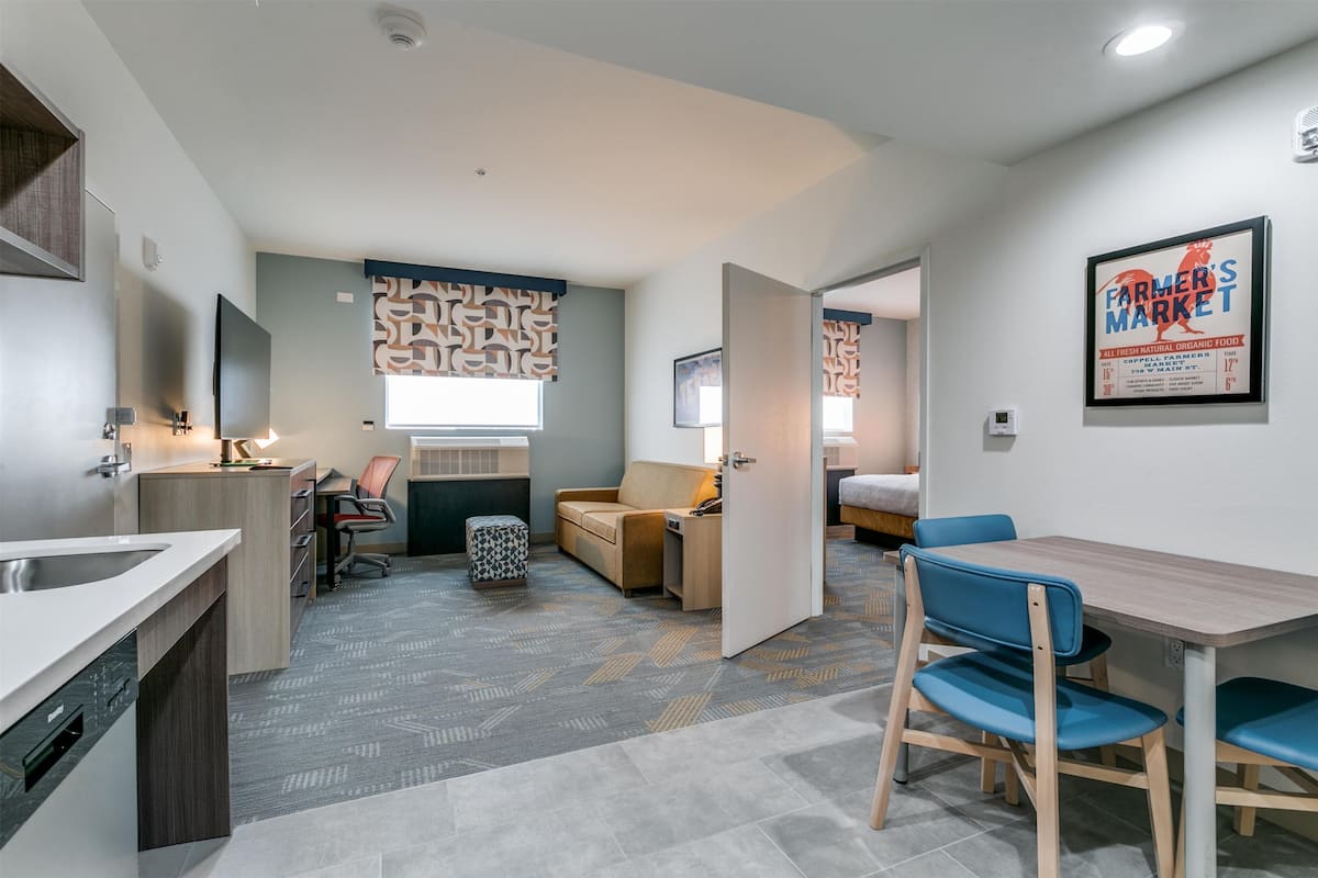 Una habitación de hotel moderna con kitchenette, un comedor con sillas azules, un sofá y un ventanal grande.