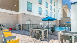 An outdoor patio with a pool, yellow chairs, a table, and a grill.