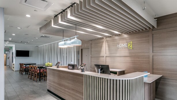 A hotel reception area with a modern design, featuring a curved reception desk, a flat-screen TV mounted on the wall, and a seating area with tables and chairs.