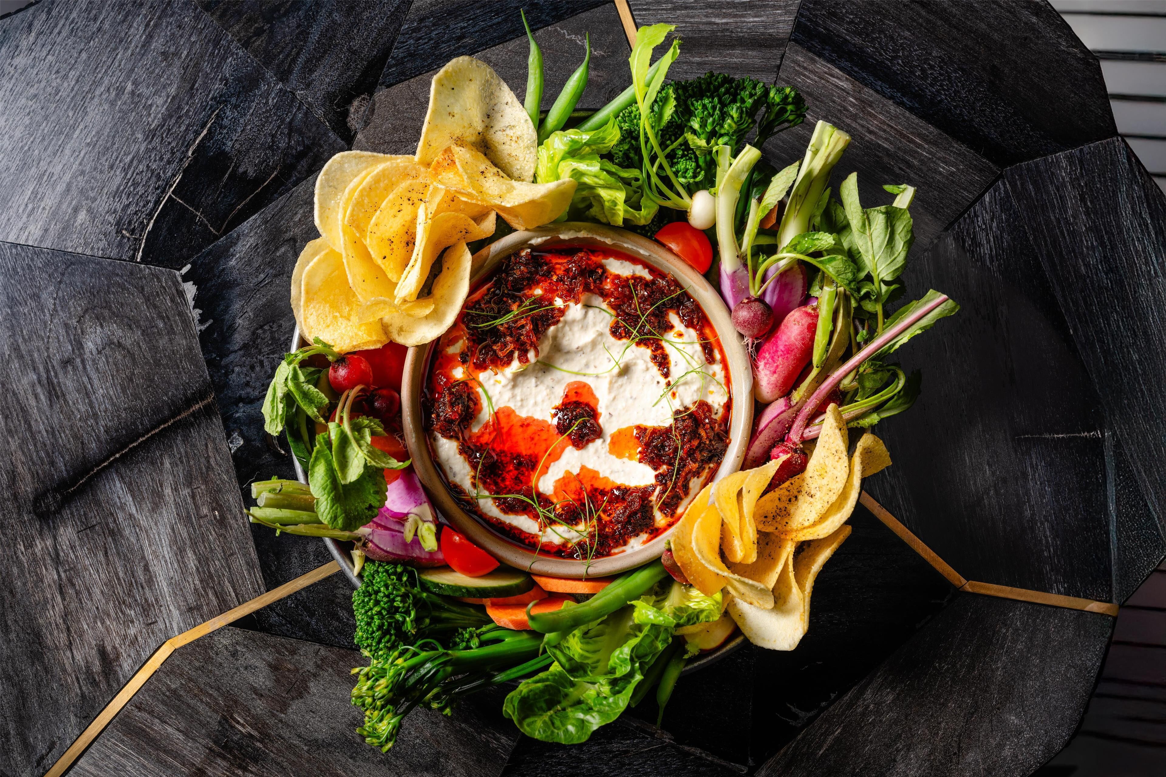 A bowl of dip surrounded by assorted vegetables and chips on a dark, textured surface.