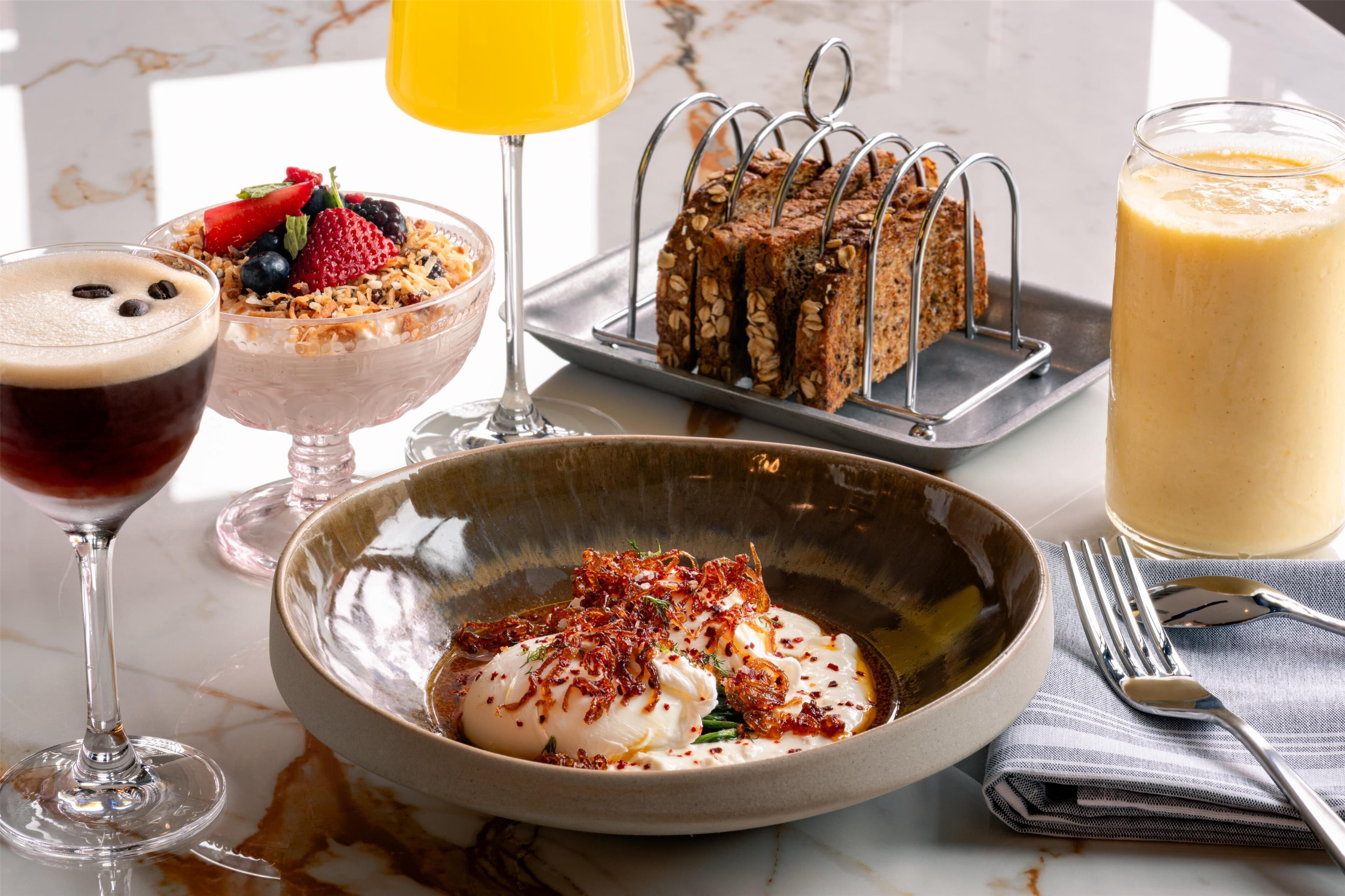 A breakfast setting with a variety of dishes including a bowl of yogurt with toppings, a fruit salad, a loaf of bread, a glass of orange juice, and a glass of milk.