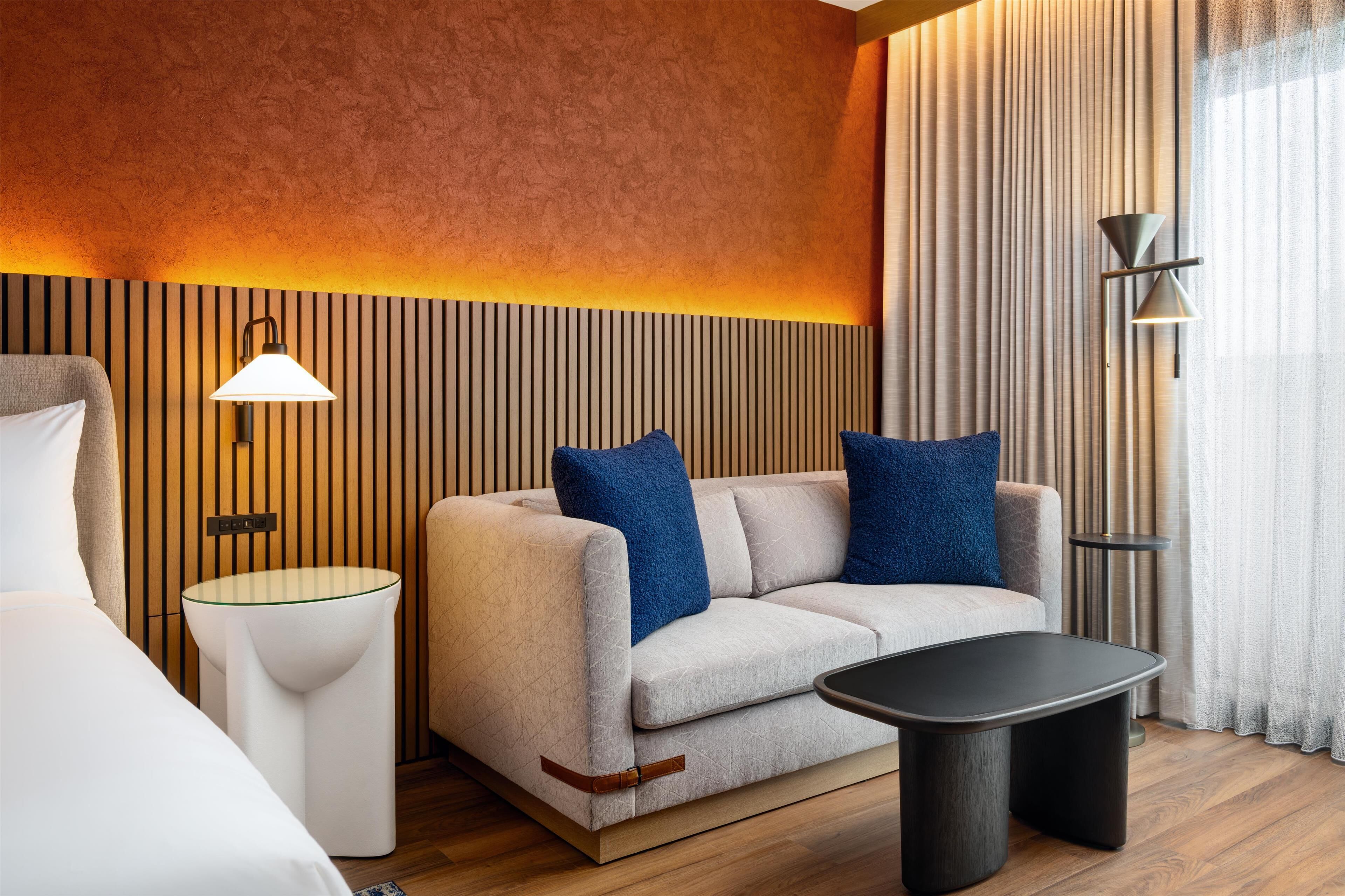 A modern hotel room with a sofa, a bedside table, a lamp, and a view of the outdoors through large windows.