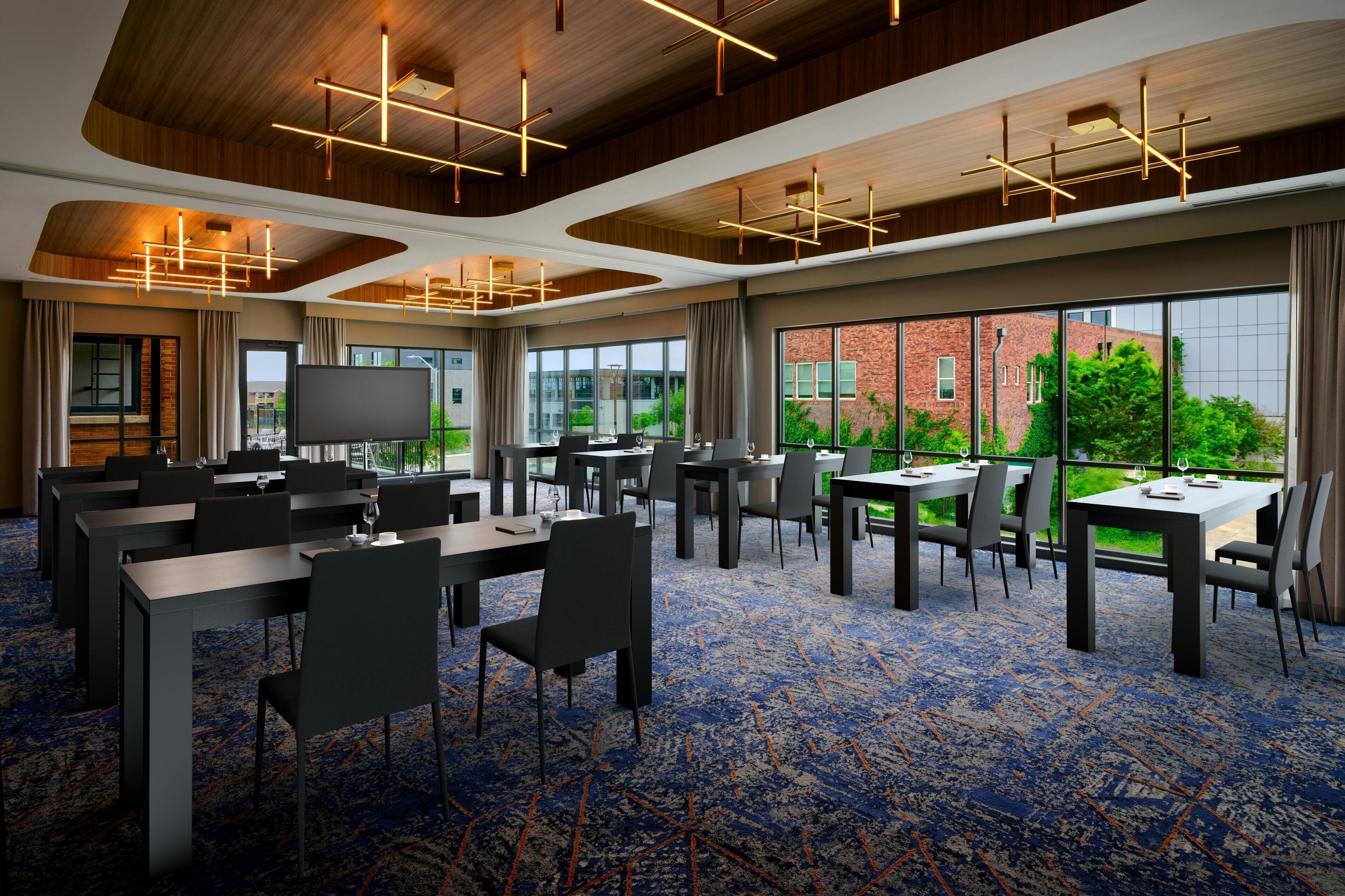 A conference room with multiple tables and chairs, large windows with a view of greenery and a brick building, and modern lighting fixtures.