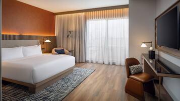 A modern hotel room with a large bed, a desk with a chair, a flat-screen TV, and a large window with curtains.