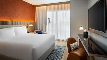 Une chambre d’hôtel avec un grand lit, un bureau doté d’un téléviseur à écran plat, une chaise et une fenêtre ornée de rideaux légers.