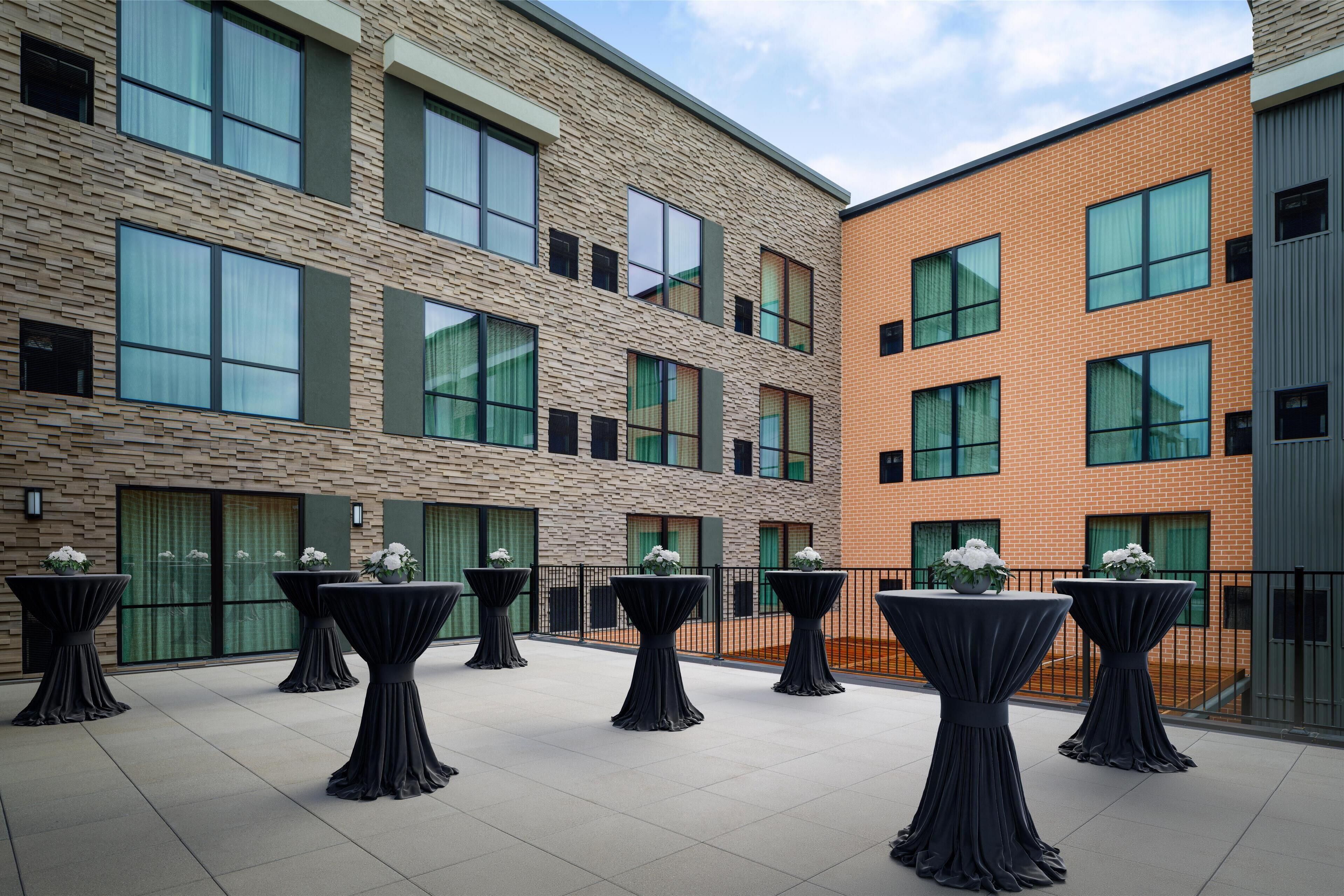 A courtyard with tables and chairs, surrounded by a building with large windows and a brick facade.